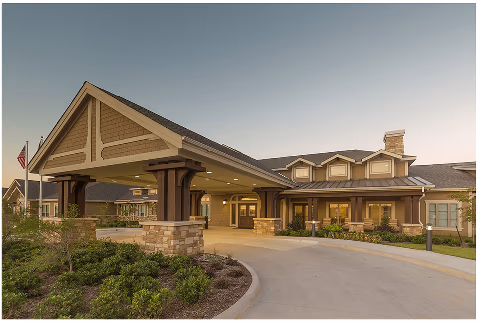 Exterior view of the Village on the Park Denton facility entrance with a covered driveway, stone and wood pillars, landscaped greenery, and an American flag on a flagpole during early evening.