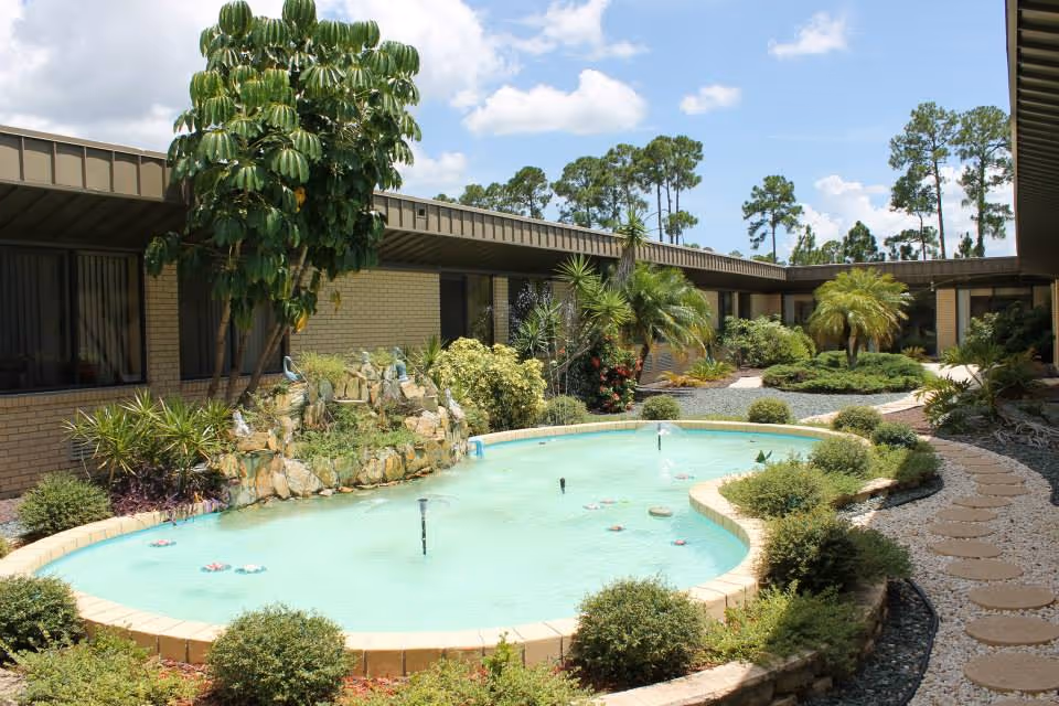 Outdoor courtyard area at North Port Rehabilitation and Nursing Center featuring a small pond with water fountains surrounded by various green plants, bushes, and trees. The courtyard is enclosed by a single-story building with windows and a covered walkway with a stone path on the right side. The sky is blue with some clouds.