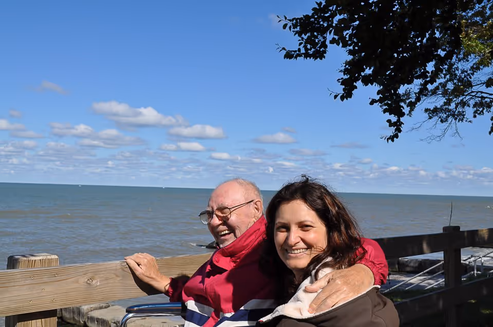 An elderly man in a wheelchair wearing a red jacket and a woman with dark hair smiling together outdoors by a wooden railing overlooking a large body of water under a blue sky with scattered clouds.