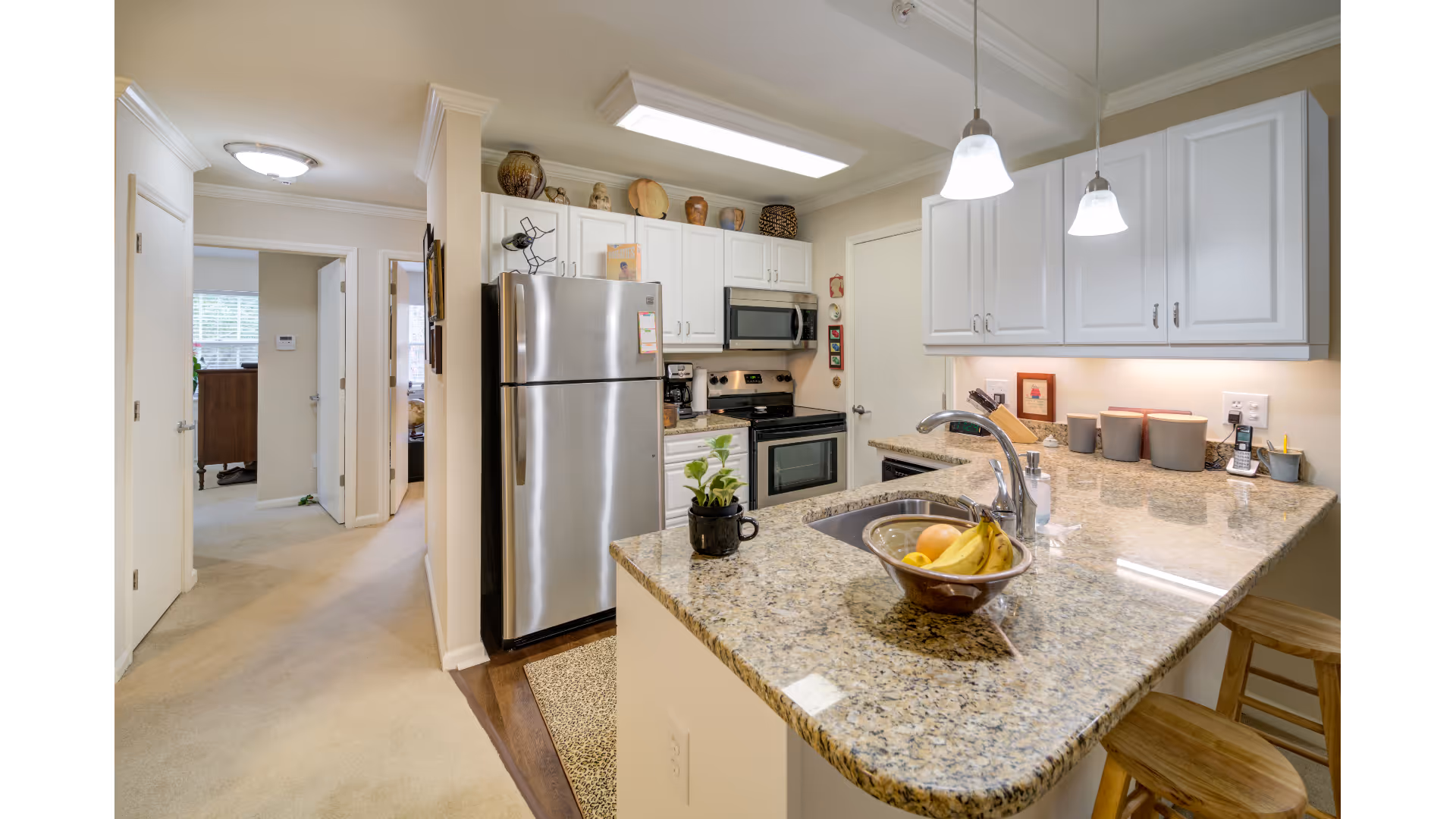 Modern kitchen with granite countertops, stainless steel refrigerator, stove, and microwave. Two pendant lights hang above the counter with a sink and a bowl of fruit. White cabinets line the walls and a hallway with carpet leads to other rooms.