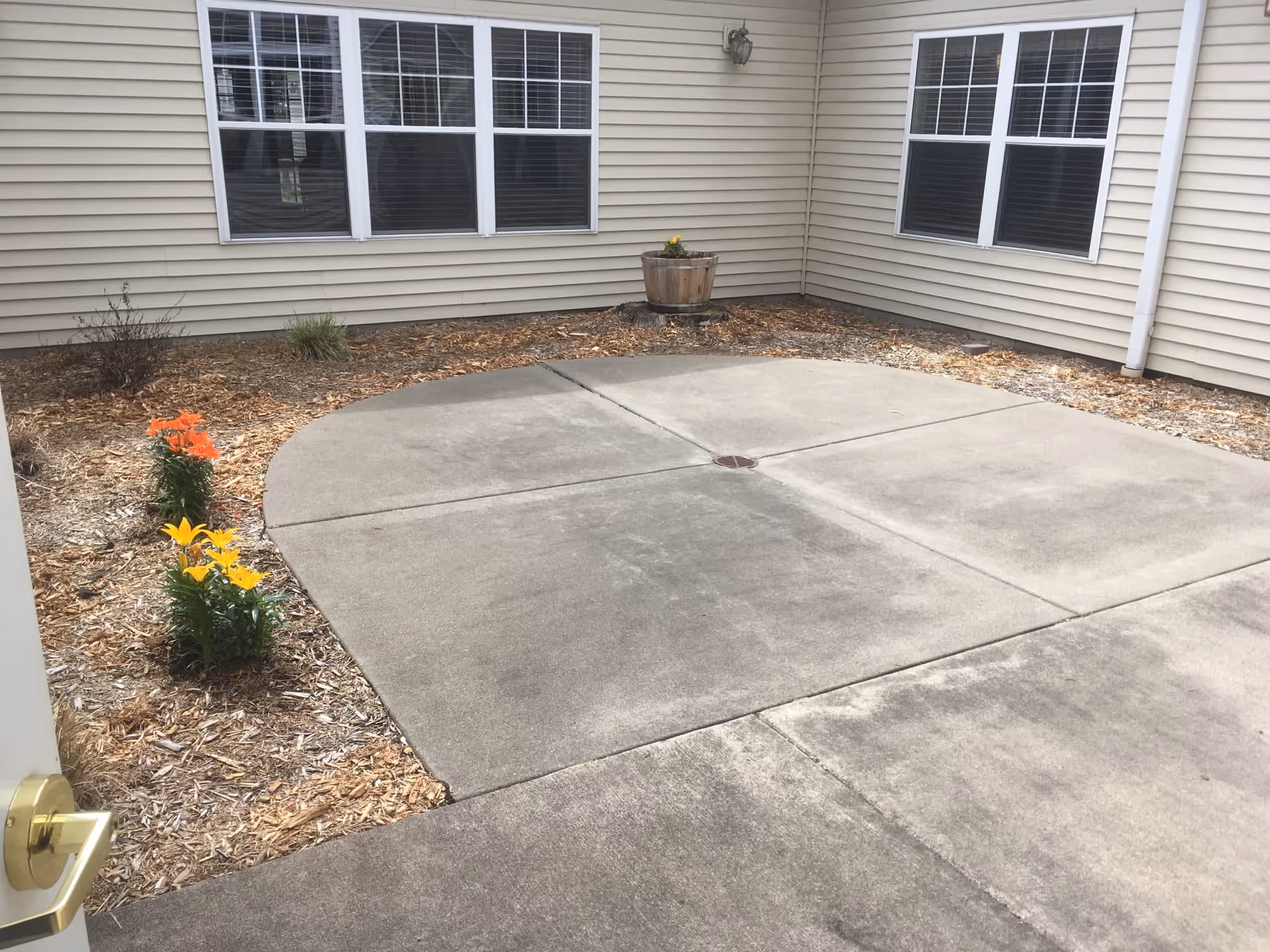 A small outdoor patio area with concrete flooring and a circular design in the center. The patio is surrounded by a garden bed with mulch and a few flowering plants, including orange and yellow flowers. The beige siding of the building and several windows with white frames are visible in the background.