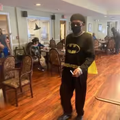 A person dressed in a Batman costume and wearing a face mask walking in a room with wooden flooring. Several other people are seated at tables and chairs in the background, engaged in various activities. The room appears to be a communal area with large windows and light-colored walls.