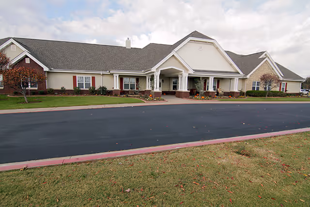Exterior view of a single-story senior living facility building with a covered entrance, beige siding, brick accents, and a dark shingled roof. The building is surrounded by a well-maintained lawn and a paved driveway in front.
