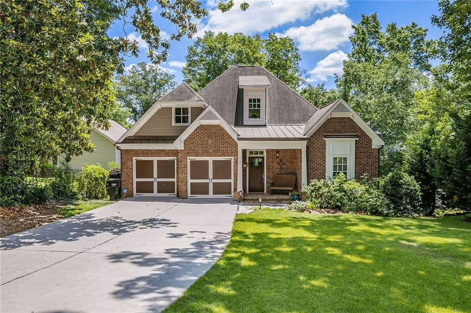 Brick single-family house with a two-car garage, front porch, driveway, and green lawn surrounded by trees.