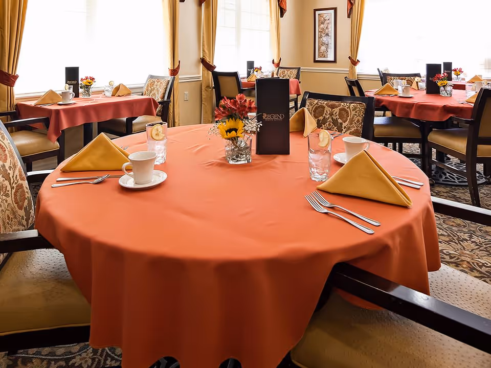 Dining room with round tables covered in orange tablecloths, each set with yellow folded napkins, cups, glasses with lemon slices, silverware, and small floral centerpieces. Chairs with patterned upholstery surround the tables. Large windows with yellow curtains allow natural light into the room.