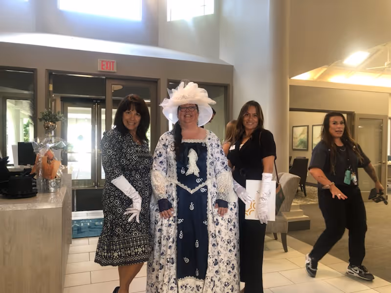 Four women standing inside a well-lit lobby area. One woman is dressed in a historical costume with a large white hat and a blue and white dress. Two other women are dressed in modern attire, one wearing a patterned dress with white gloves and the other in black holding a folder and also wearing white gloves. The fourth woman is casually dressed and walking in the background. The lobby has large windows, a reception desk, and seating areas.