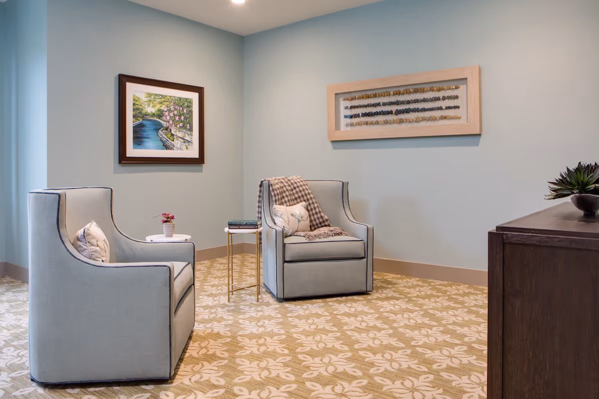 Two upholstered armchairs and a small side table in a softly lit lounge area with framed artwork on the walls.