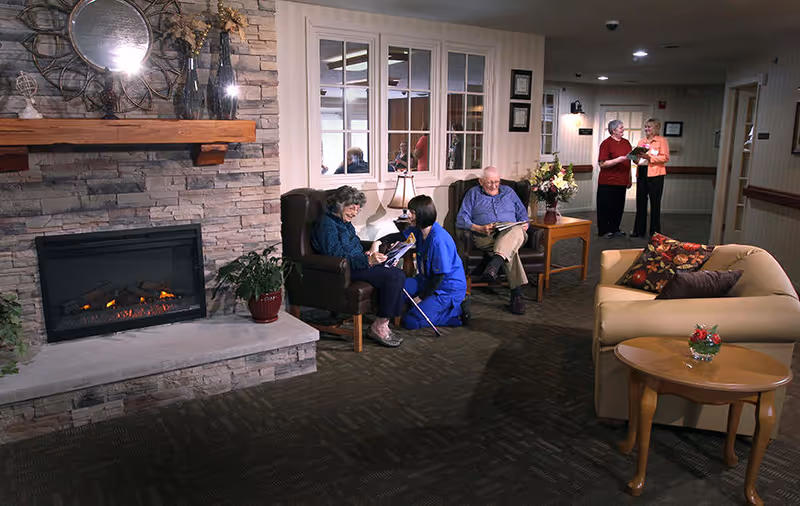 A cozy common area in a senior living facility with a stone fireplace on the left, a woman sitting in a chair talking to a caregiver kneeling beside her, an elderly man sitting on a chair reading a newspaper, and two women standing and conversing in the background. The room has a beige couch with floral pillows and a wooden side table with a small plant.