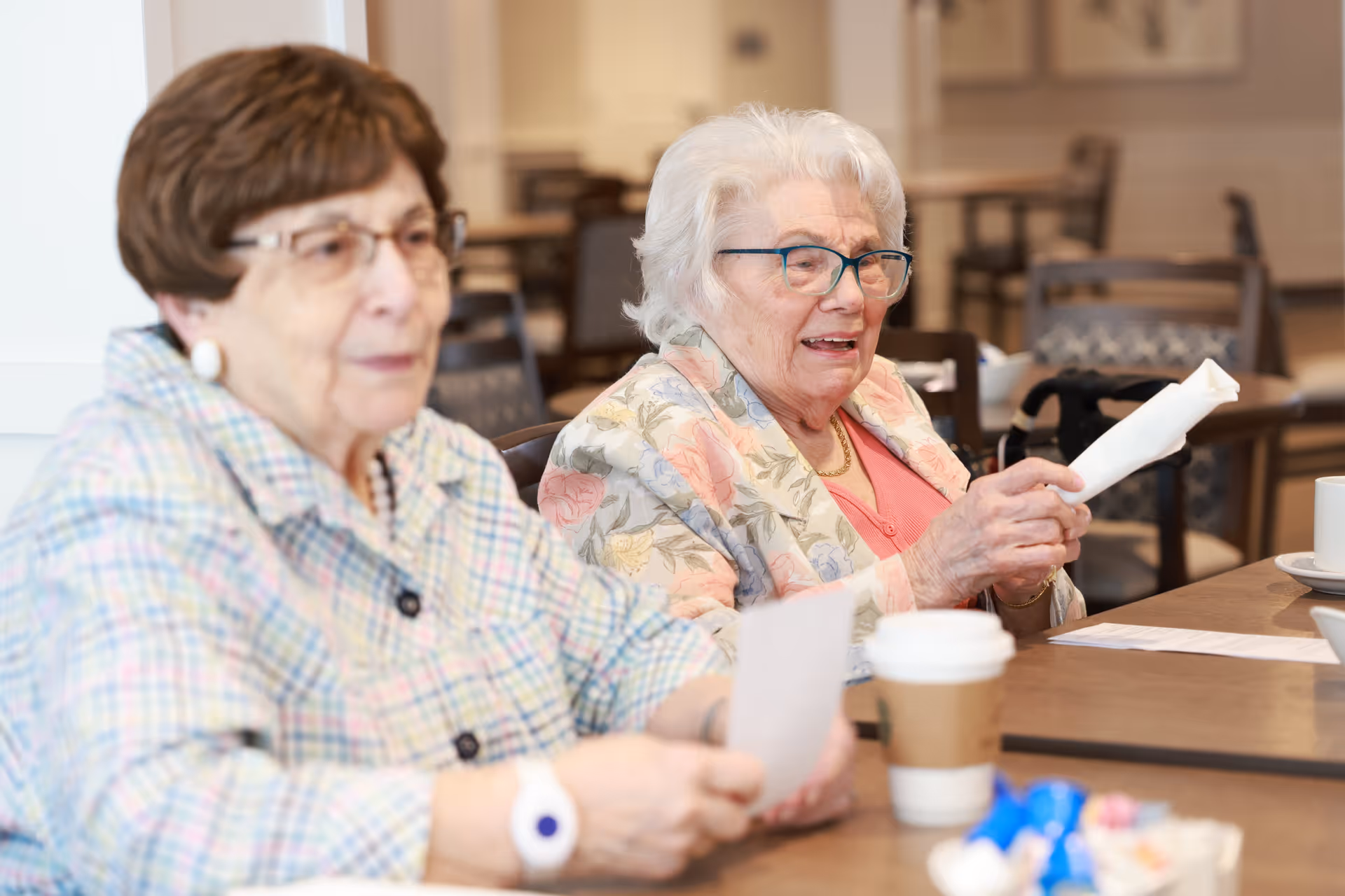Two elderly women sitting at a table in a communal dining area, one holding a rolled napkin and the other holding a piece of paper, with coffee cups and other dining items on the table.