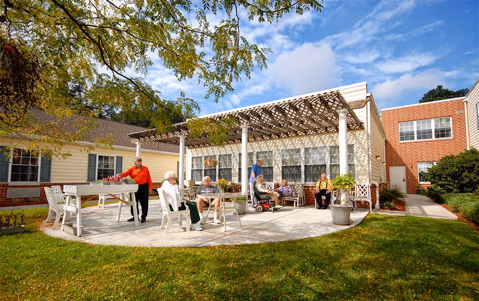 Outdoor patio area at Chestnut Knoll with several elderly people sitting and standing under a pergola attached to a building. The patio is surrounded by green grass and trees, with a partly cloudy sky overhead.