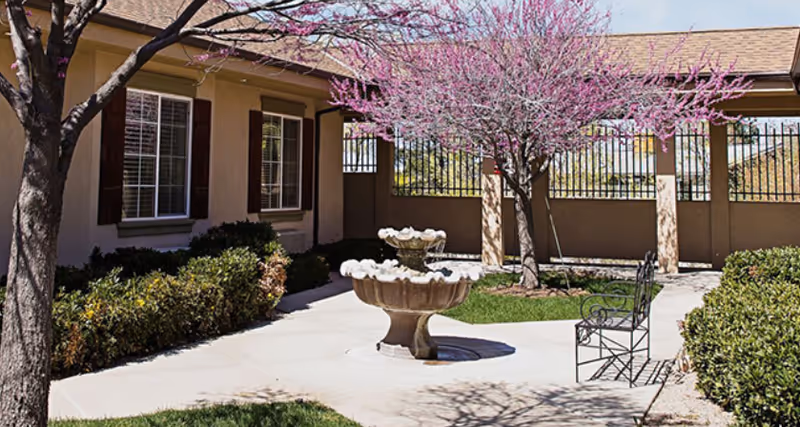 Outdoor courtyard area at Alta Ridge Assisted Living of Holladay featuring a two-tiered stone fountain in the center, surrounded by a paved walkway, green bushes, and a tree with pink blossoms. There is a metal bench on the right side and the building with windows and brown shutters is visible in the background.