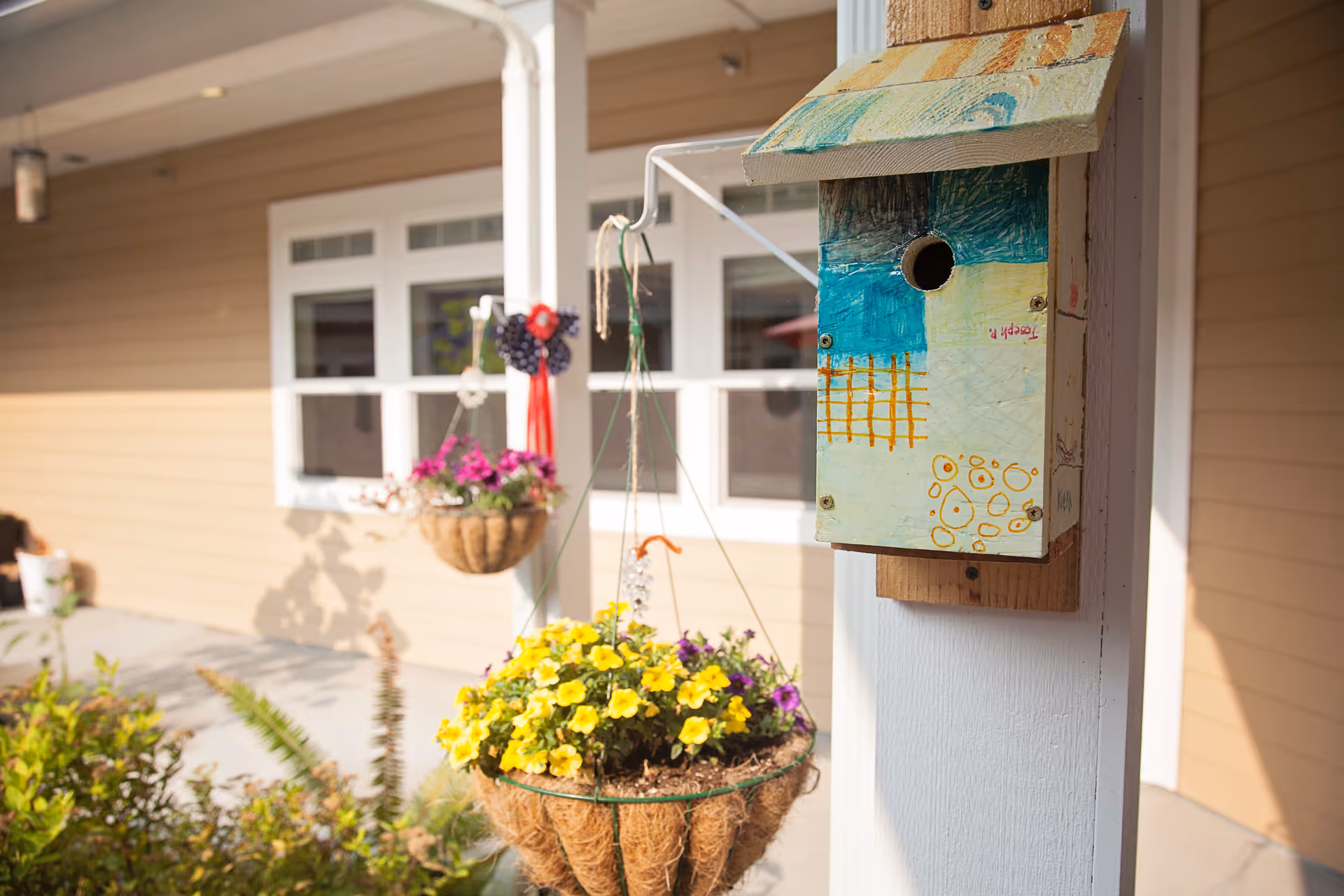 A painted birdhouse mounted on a porch post next to hanging flower baskets in front of a beige-sided building.