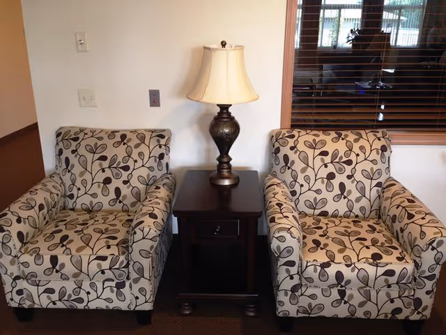 Two beige armchairs with a dark brown leaf pattern are positioned on either side of a small dark wooden side table with a drawer. On the table is a decorative lamp with a beige lampshade. Behind the chairs is a white wall with electrical outlets and a window with wooden blinds showing a reflection of another room.