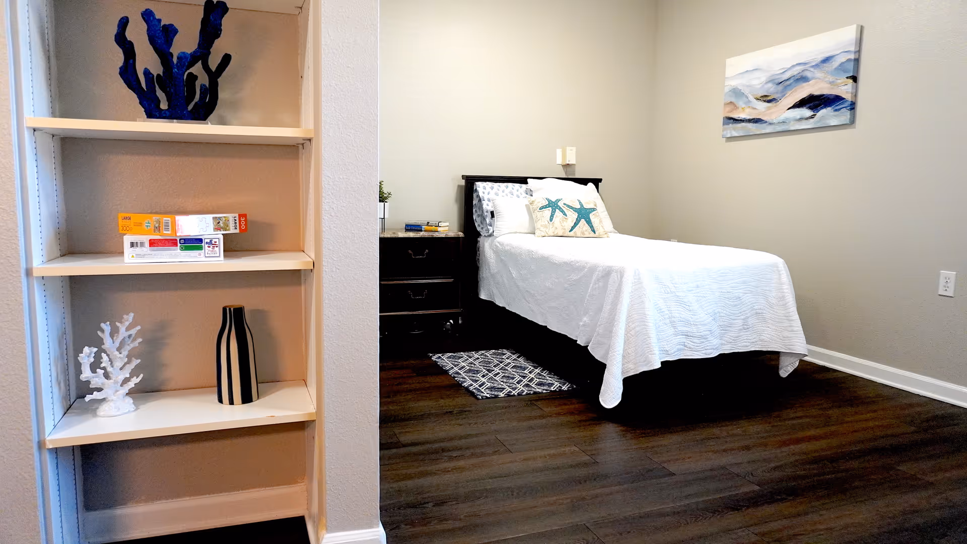 A simple bedroom with a single bed covered in a white quilt and two decorative pillows with starfish designs. Next to the bed is a dark wooden nightstand with books and a small plant. On the wall above the bed is a framed abstract painting with blue and beige tones. To the left, there is a built-in shelving unit with decorative items including a blue coral sculpture, a white coral sculpture, a striped vase, and some board games. The floor is dark wood, and the walls are painted light beige.