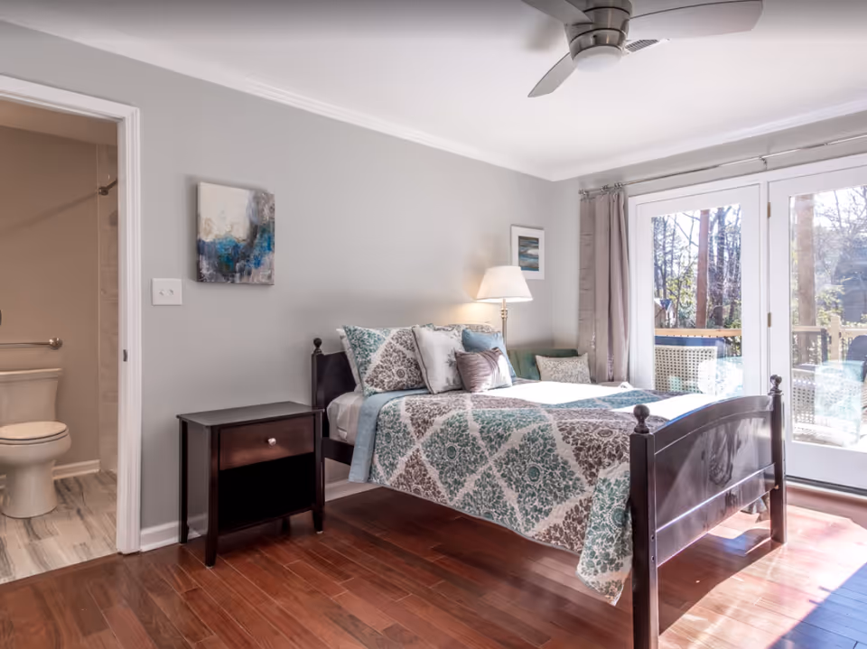 Sunlit bedroom with a wooden bed dressed in patterned bedding, a nightstand and lamp, sliding glass doors to a deck, and an open door showing an en suite bathroom.