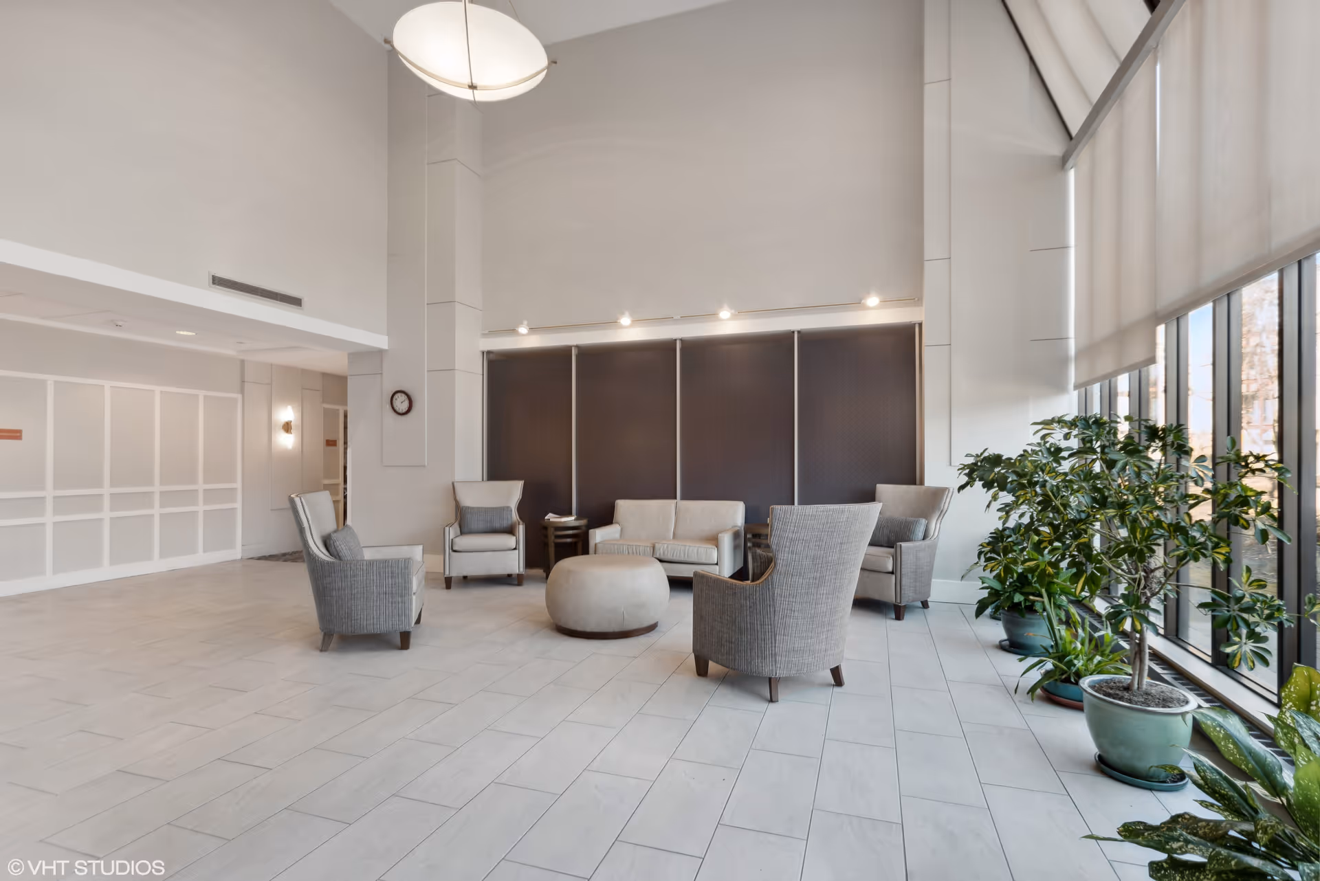 A spacious, modern common area with high ceilings and large windows letting in natural light. The room features a seating arrangement with a sofa, four armchairs, a round ottoman, and side tables. Several potted plants are placed near the windows, and a clock is mounted on a column. The floor is tiled in light colors, and the walls are painted white.