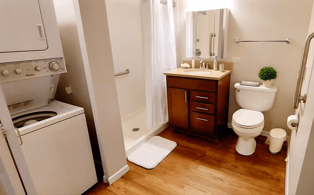 A bathroom with a wooden floor featuring a stacked washer and dryer on the left, a walk-in shower with a white curtain, a wooden vanity with a sink and mirror, a toilet with a small green plant and folded towels on top, and grab bars on the walls for accessibility.