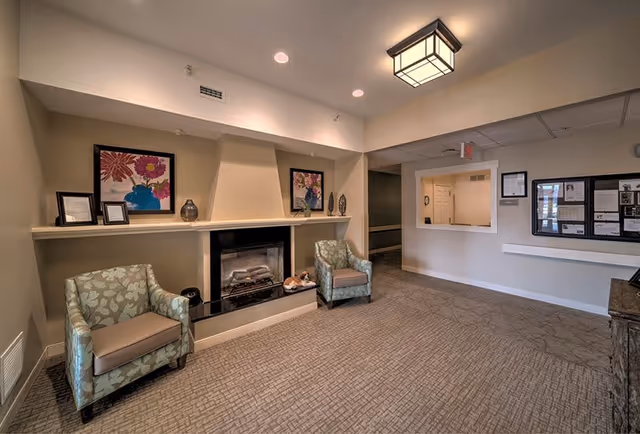 A cozy senior living facility common area with two patterned armchairs placed on either side of a fireplace. Above the fireplace are decorative items including framed floral artwork and small sculptures. The room has beige walls, carpeted floor, recessed lighting, and a square ceiling light fixture. There is a reception window and bulletin boards on the far wall.