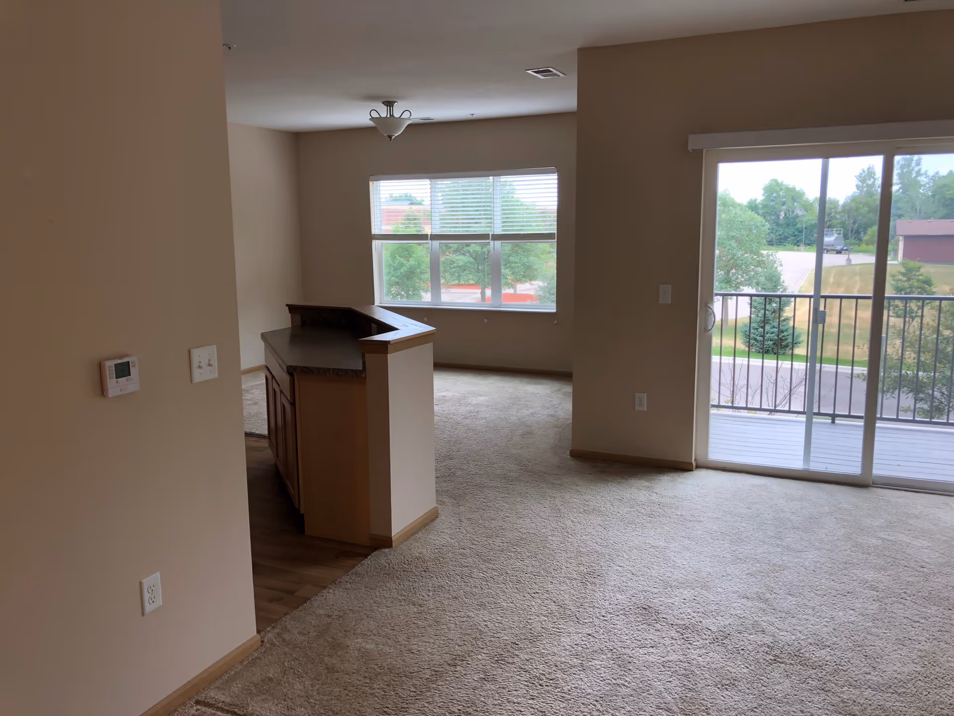 Interior view of an empty senior living apartment with beige walls and carpeted floors. There is a small kitchen counter with wooden cabinets on the left side, a large window in the background, and a sliding glass door leading to a balcony with a view of trees and a parking area outside.
