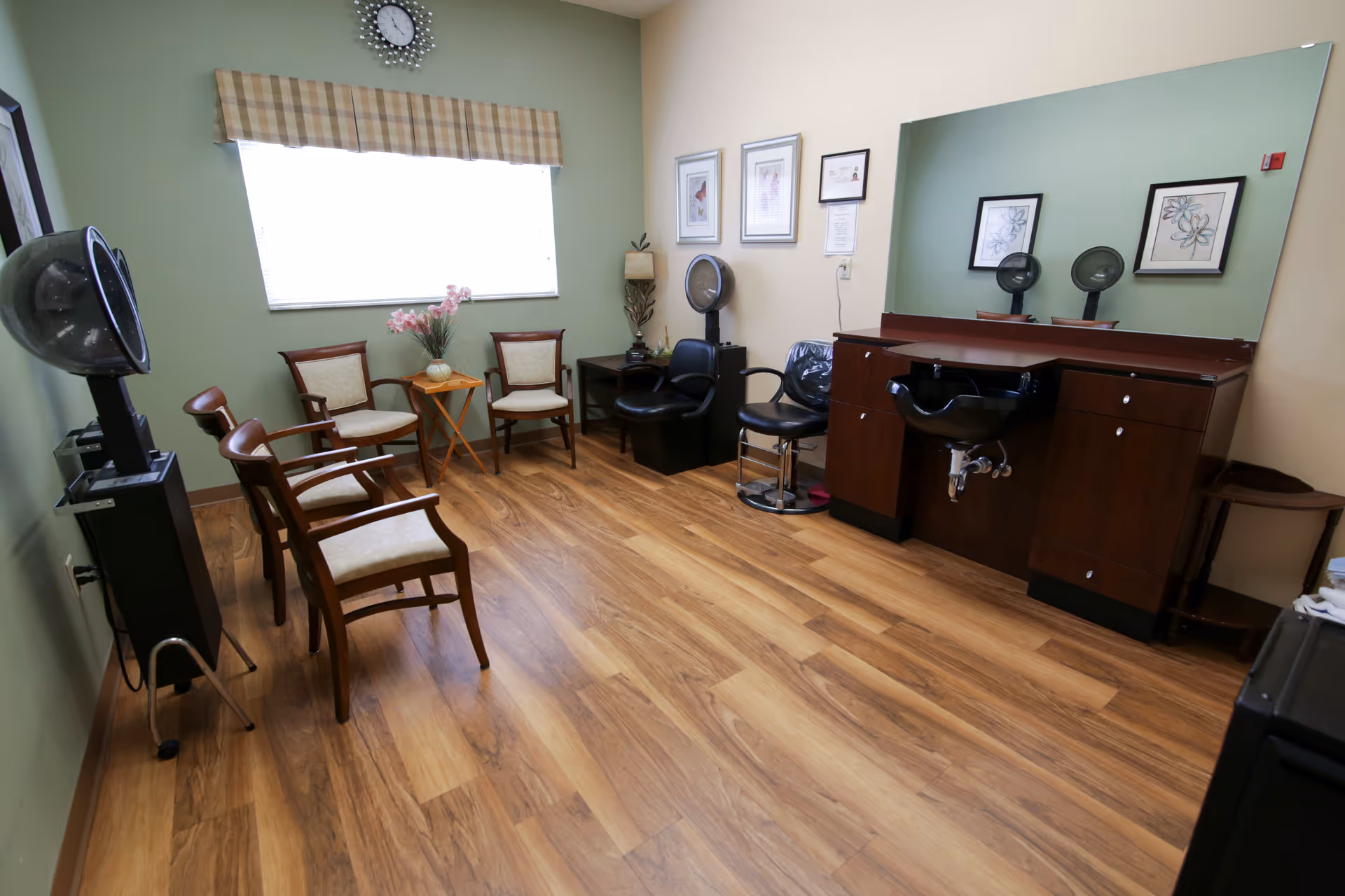 A bright salon-style room with wood floors, several seating chairs, hair dryers, and a styling station with a sink and large mirror.