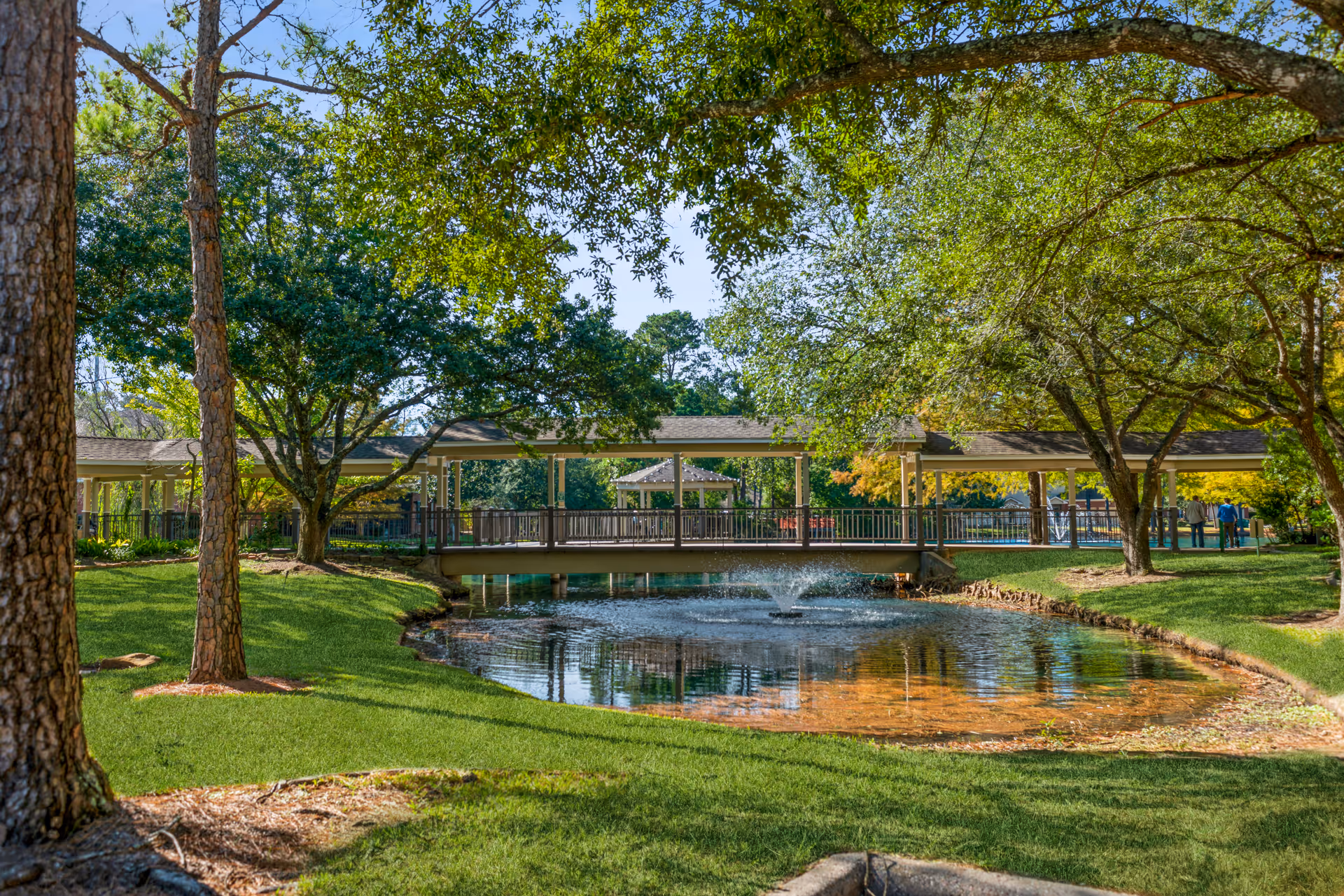 A peaceful outdoor scene at The Forum at The Woodlands featuring a small pond with a water fountain in the center, surrounded by green grass and large trees. A covered wooden bridge spans the pond, and a gazebo is visible in the background. The area is shaded by the trees, creating a serene and inviting atmosphere.