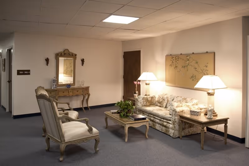 A cozy sitting area in a hallway with a floral-patterned sofa, two upholstered armchairs, a wooden coffee table with a plant and books, two table lamps on side tables, a decorative wall mirror above a wooden console table, and soft lighting creating a warm ambiance.