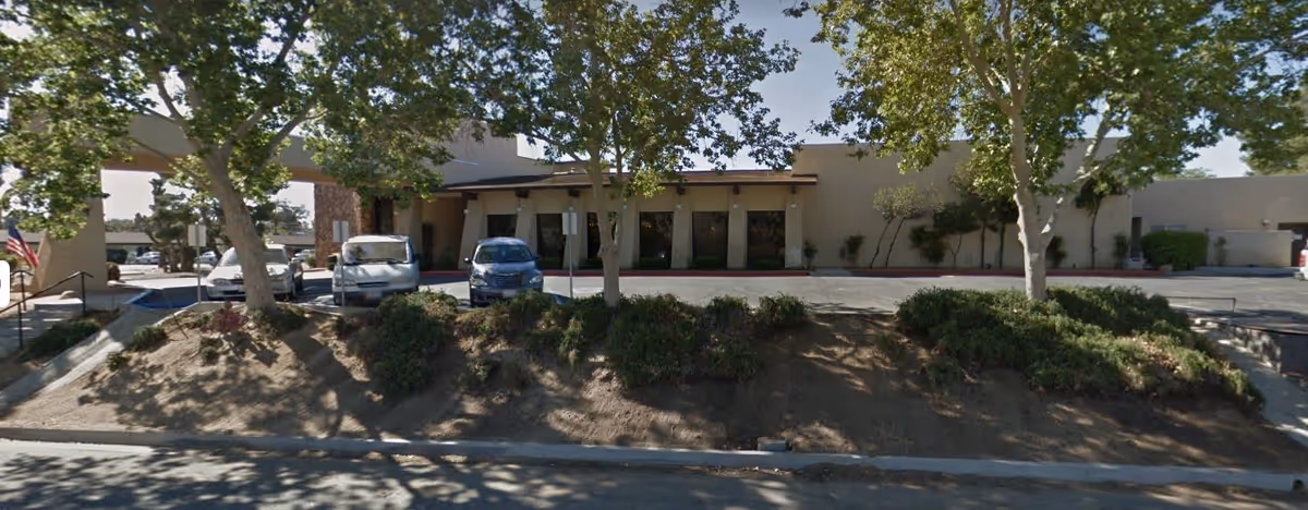 Exterior view of Santa Fe Assisted Living facility showing a beige building with several windows, a covered entrance, and a parking area with cars. There are trees and shrubs in front of the building casting shadows on the ground.