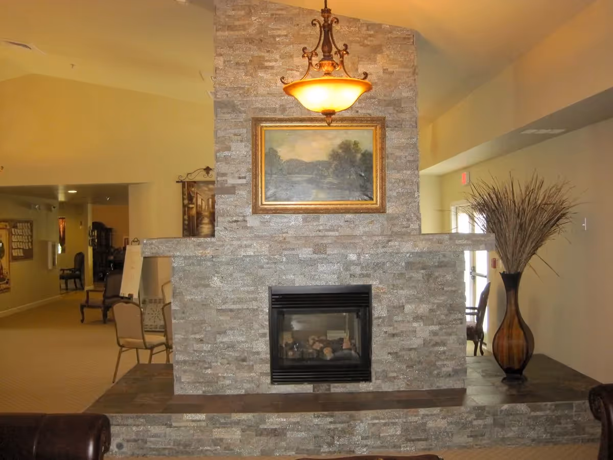 Interior view of a senior living facility lounge area featuring a stone fireplace with a framed landscape painting above it. A decorative hanging light fixture is centered above the fireplace. To the right, there is a large vase with tall dried plants. Chairs and tables are visible in the background along a hallway.