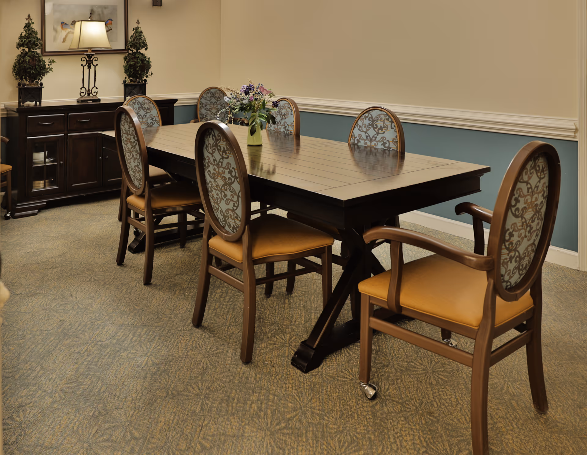 A long wooden dining table with six upholstered chairs and a floral centerpiece in a softly decorated dining room.