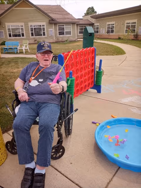An elderly man in a wheelchair holding a pink fishing pole, playing a fishing game outside on a paved area. Behind him is a large red and blue Connect Four game set up on the sidewalk. There is a blue plastic kiddie pool with colorful floating toy fish nearby. The background shows a building with beige siding and multiple windows, along with some green grass and outdoor benches.
