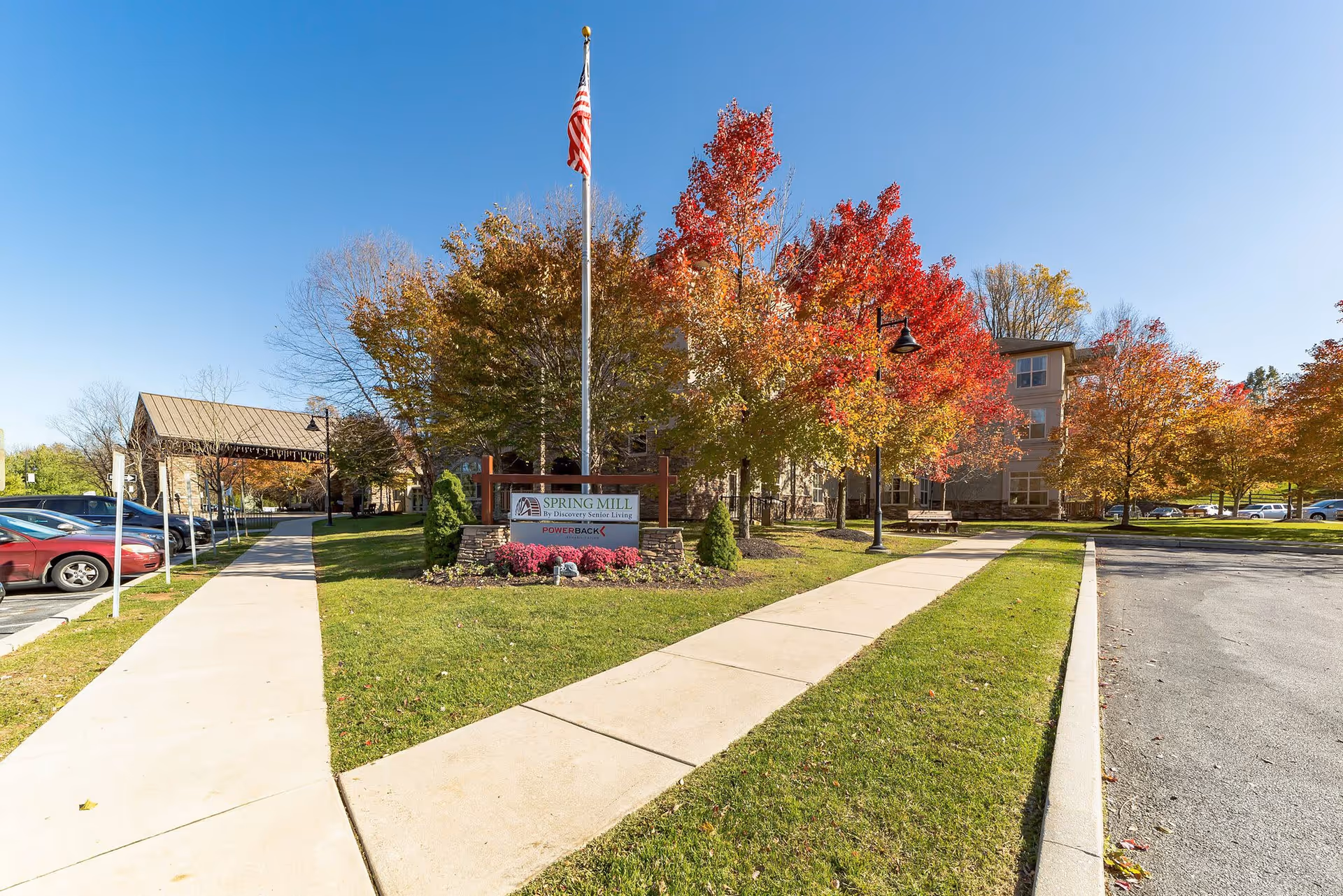 Exterior view of Spring Mill senior living facility on a clear day with blue sky. The scene shows a paved walkway leading to the entrance, surrounded by green grass and trees with autumn foliage in shades of red, orange, and yellow. An American flag is flying on a flagpole near a sign that reads 'Spring Mill by Discovery Senior Living POWERBACK'. Several parked cars are visible on the left side.