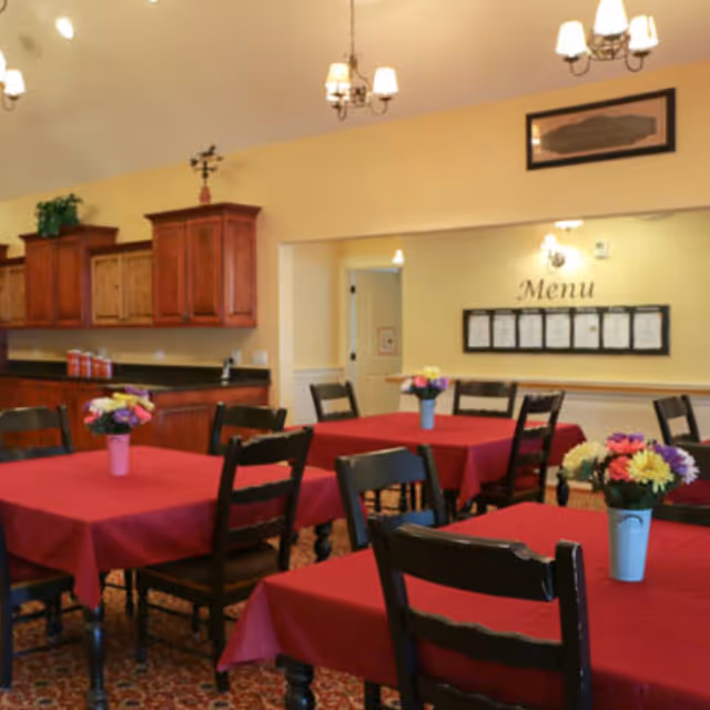 Dining room with multiple tables covered in red tablecloths, each table decorated with a small vase of colorful flowers. Wooden chairs surround the tables. The room has warm yellow walls, wooden cabinets along one side, and a menu displayed on the far wall under soft lighting fixtures.