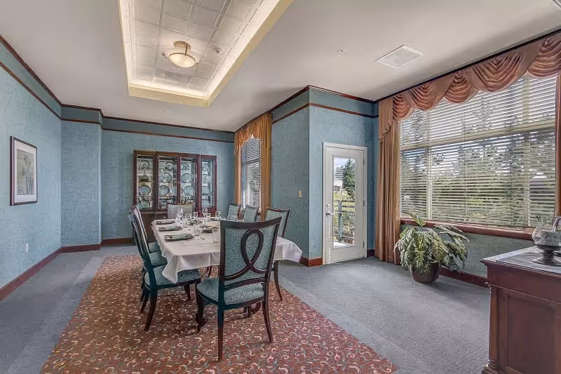 A formal dining room with a long table set for a meal, surrounded by eight chairs. The room has blue walls, a patterned carpet, large windows with drapes, and a glass door leading outside. A china cabinet with dishes is against one wall, and a potted plant sits near the window.