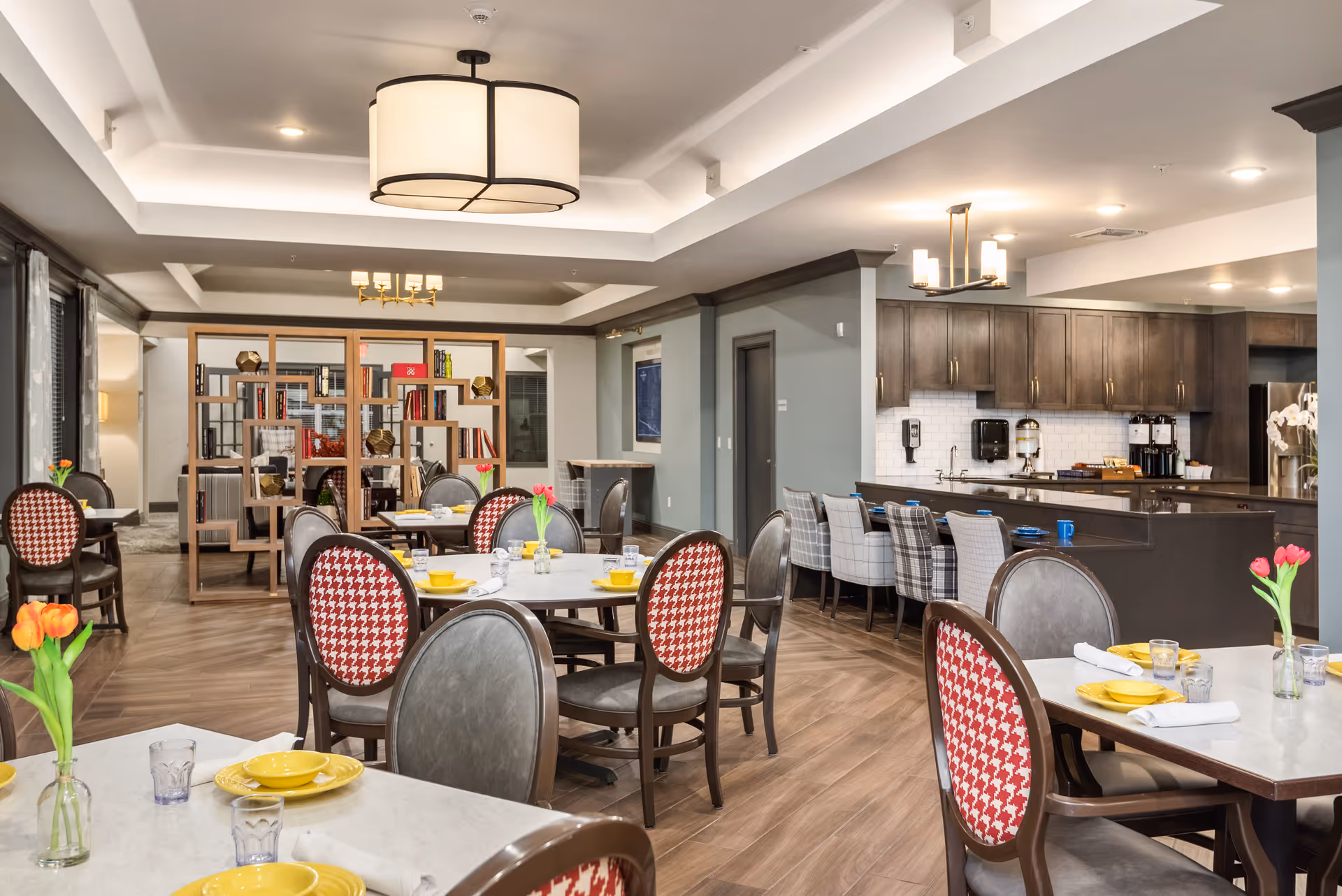 A bright and modern dining area in a senior living facility with several tables set with yellow plates, glasses, and white napkins. The chairs have a mix of red and white houndstooth patterned backs and solid gray backs. There are fresh tulip flowers in small vases on the tables. In the background, there is a kitchen area with dark wood cabinets, a coffee station, and a counter with plaid upholstered bar stools. A wooden bookshelf with decorative items and books separates the dining area from another room.