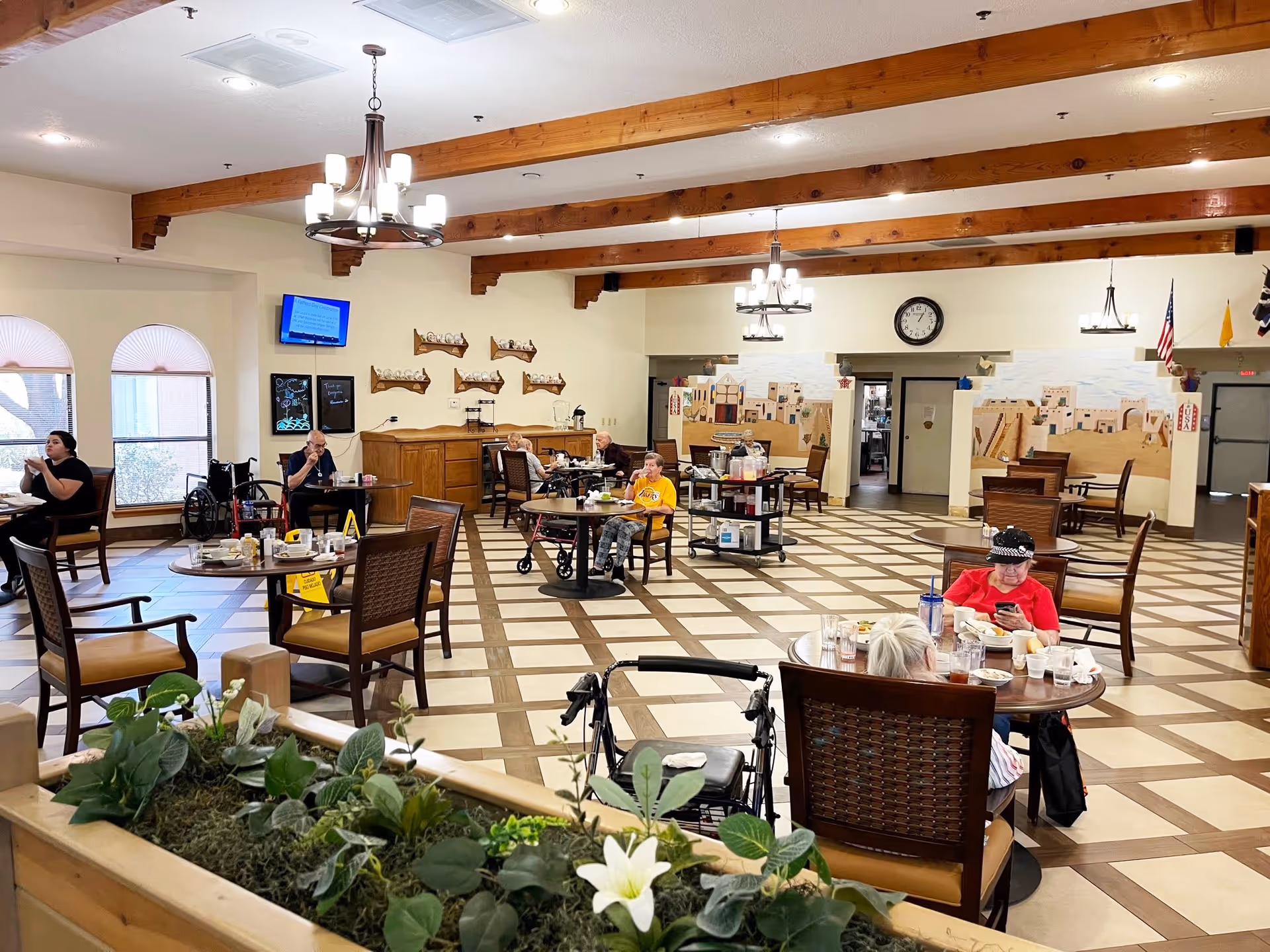 A spacious dining room in an assisted living facility with several elderly residents seated at round tables eating and socializing. The room features wooden ceiling beams, chandeliers, large windows with arched blinds, and a tiled floor with a checkered pattern. There are decorative murals on the walls and a clock above a doorway. A plant arrangement is visible in the foreground.
