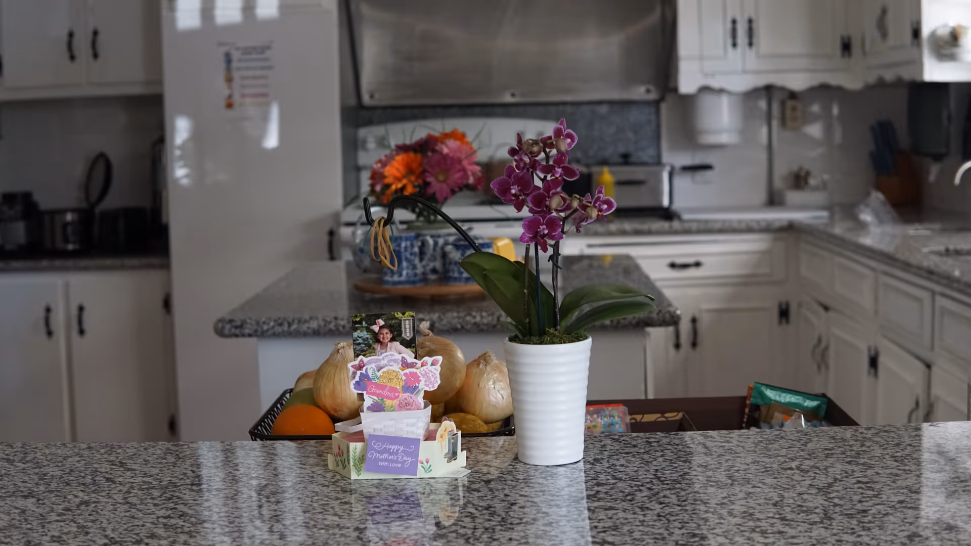 A kitchen interior with white cabinets and granite countertops. On the countertop in the foreground, there is a white pot with a purple orchid plant, a Mother's Day card, and a basket containing onions and other fruits. In the background, there is a refrigerator, stove, and various kitchen items.