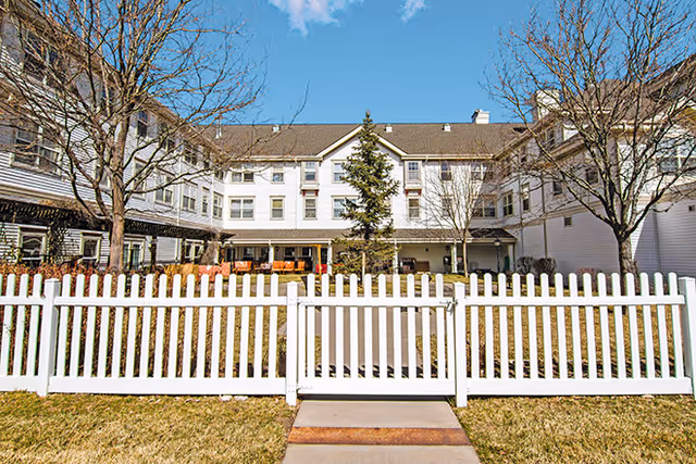 View of a senior living facility building named Woodland Estates Senior Living with a white picket fence in the foreground, leafless trees, and a clear blue sky.