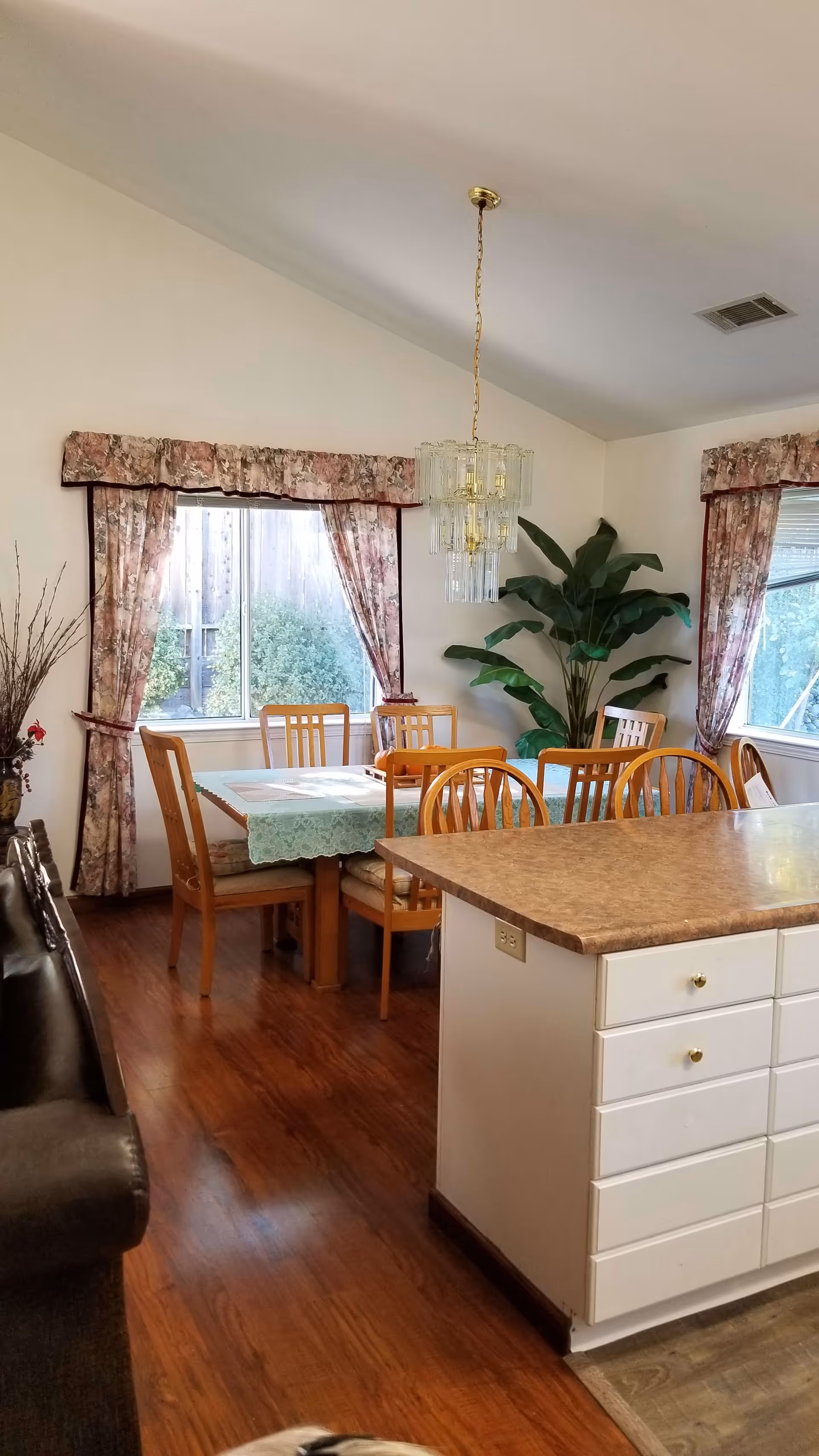 Open dining area and kitchen island with a wooden table and chairs, chandelier, large plant, and floral curtains.