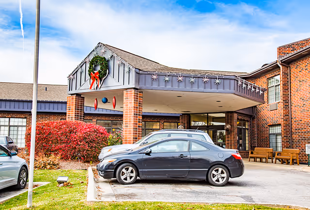 Front entrance of a brick building decorated with a large green wreath with a red bow and hanging ornaments under a covered driveway. Several cars are parked in front, and there are benches near the entrance. The sky is partly cloudy.