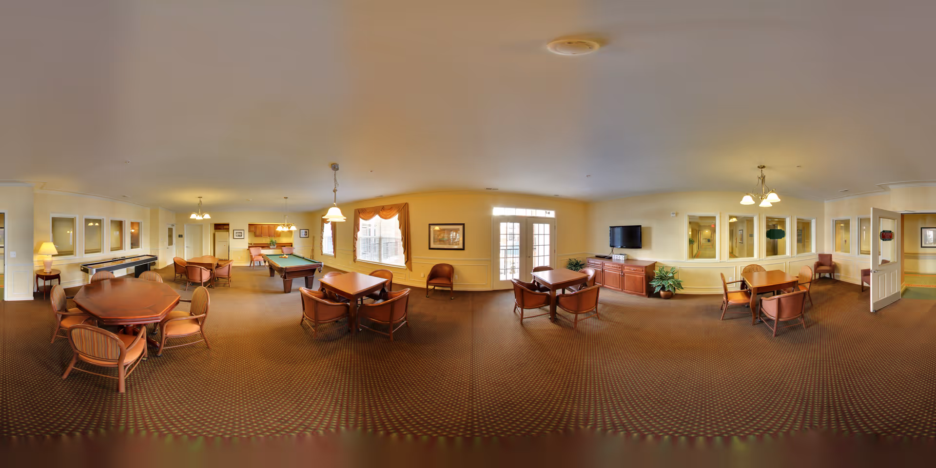 A spacious common area in a senior living facility with multiple tables and chairs arranged for socializing or games. The room features a pool table, a shuffleboard table, a television mounted on a wooden cabinet, and large windows with curtains allowing natural light to enter. The walls are painted a warm yellow, and the carpet has a patterned design. Several light fixtures hang from the ceiling, and there are plants placed around the room for decoration.