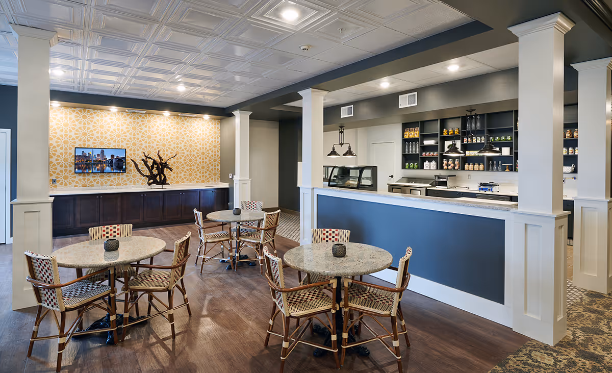 Community dining area with round tables and wicker chairs facing a service counter and shelving against a decorative wall.