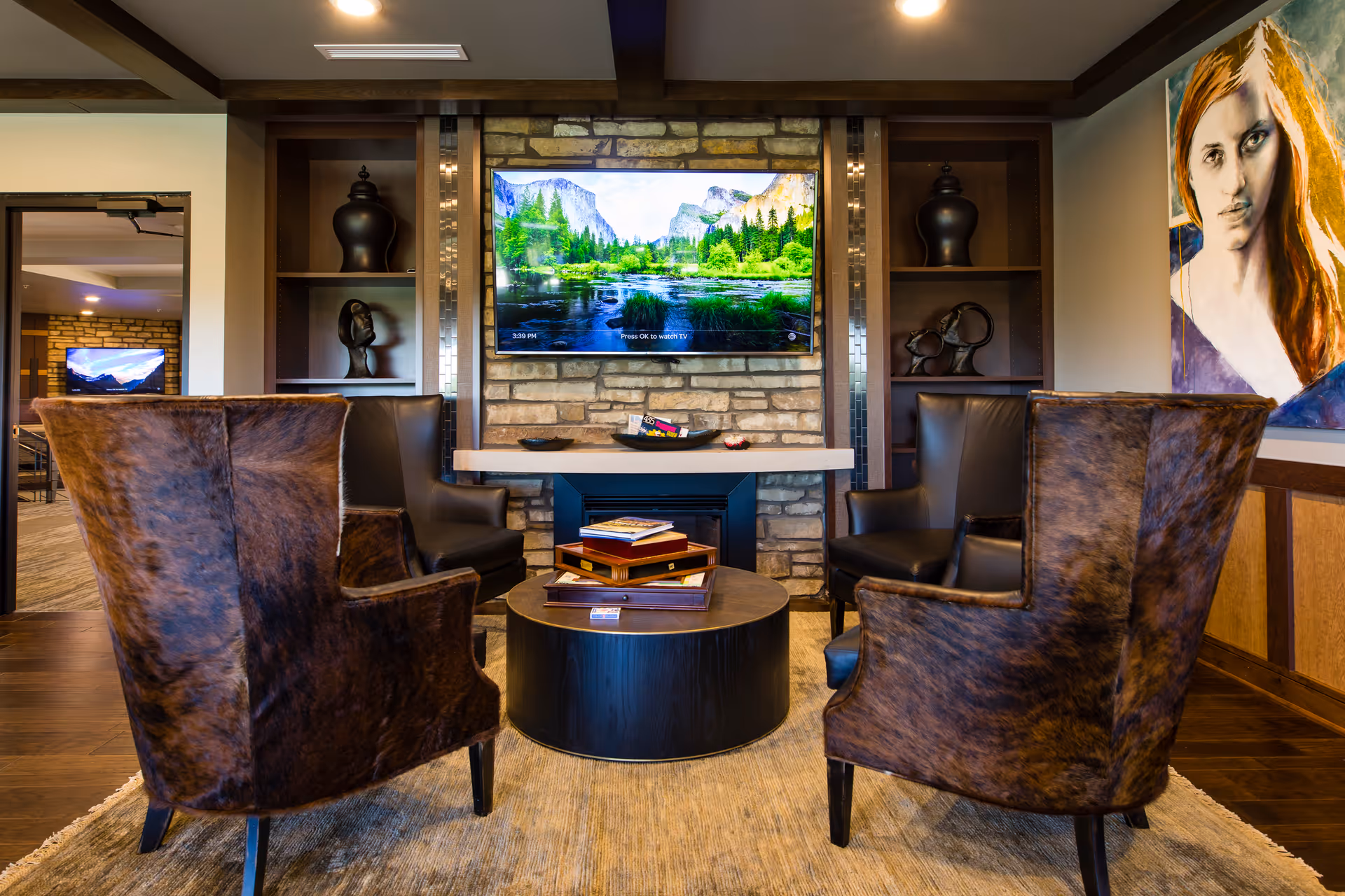 A cozy living room area with four brown leather armchairs arranged around a round black coffee table with books on it. A flat-screen TV is mounted on a stone wall above a modern fireplace. Built-in shelves with decorative items flank the TV. A large portrait painting of a woman is on the right wall.