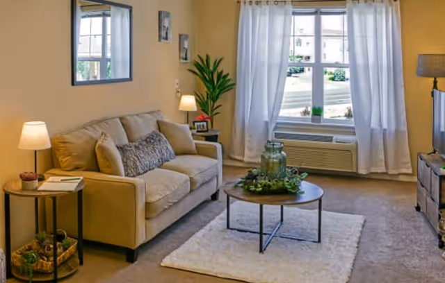 A cozy living room with a beige sofa adorned with cushions, a round wooden coffee table on a white rug, a side table with a lamp and books, a tall green plant in the corner, and a large window with white curtains letting in natural light.