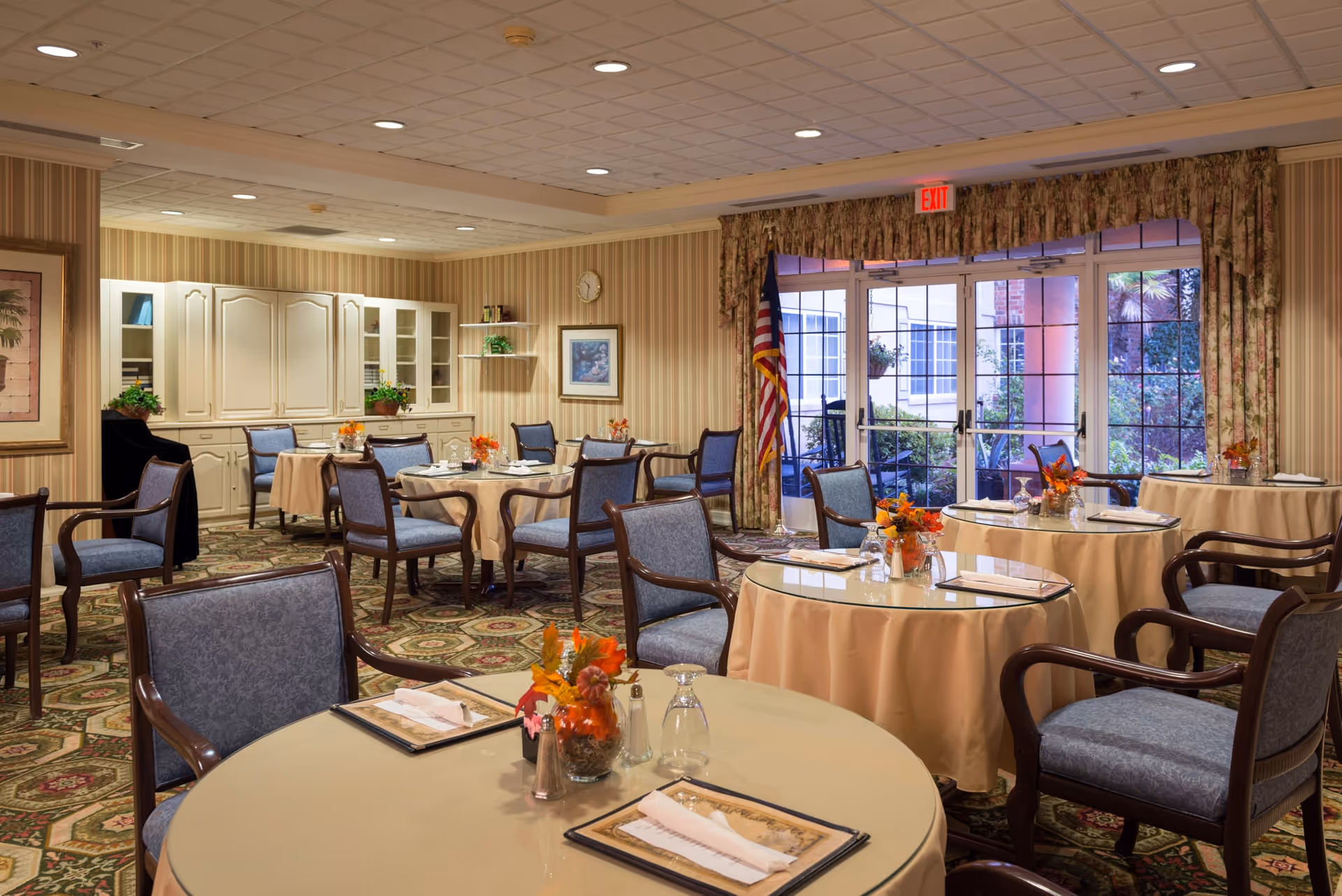 A dining room with round tables covered in beige tablecloths and set with menus, napkins, glasses, and small floral centerpieces. The room has patterned carpet, striped wallpaper, and a cabinet with shelves holding plants and books. Large windows and glass doors with floral curtains lead to an outdoor area. An American flag stands near the exit door.