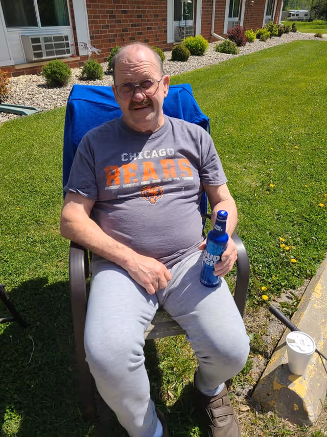 An elderly man wearing glasses, a Chicago Bears t-shirt, and gray sweatpants is sitting outdoors on a chair with a blue towel draped over the back. He is holding a blue Bud Light bottle in his right hand. Behind him is a brick building with windows and a well-maintained lawn with small bushes and rocks along the building's edge. A white disposable cup with a lid is on the ground near the curb beside him.