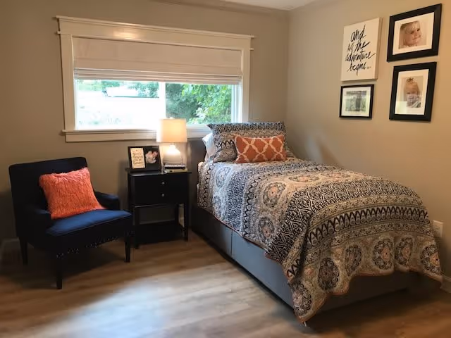 A cozy bedroom with a single bed covered in a patterned quilt and decorative pillows. Next to the bed is a black nightstand with a lamp, framed photos, and a small book. A navy blue armchair with an orange pillow is positioned to the left of the nightstand. The room has light-colored walls, a large window with a white blind, and framed pictures hanging on the wall above the bed.