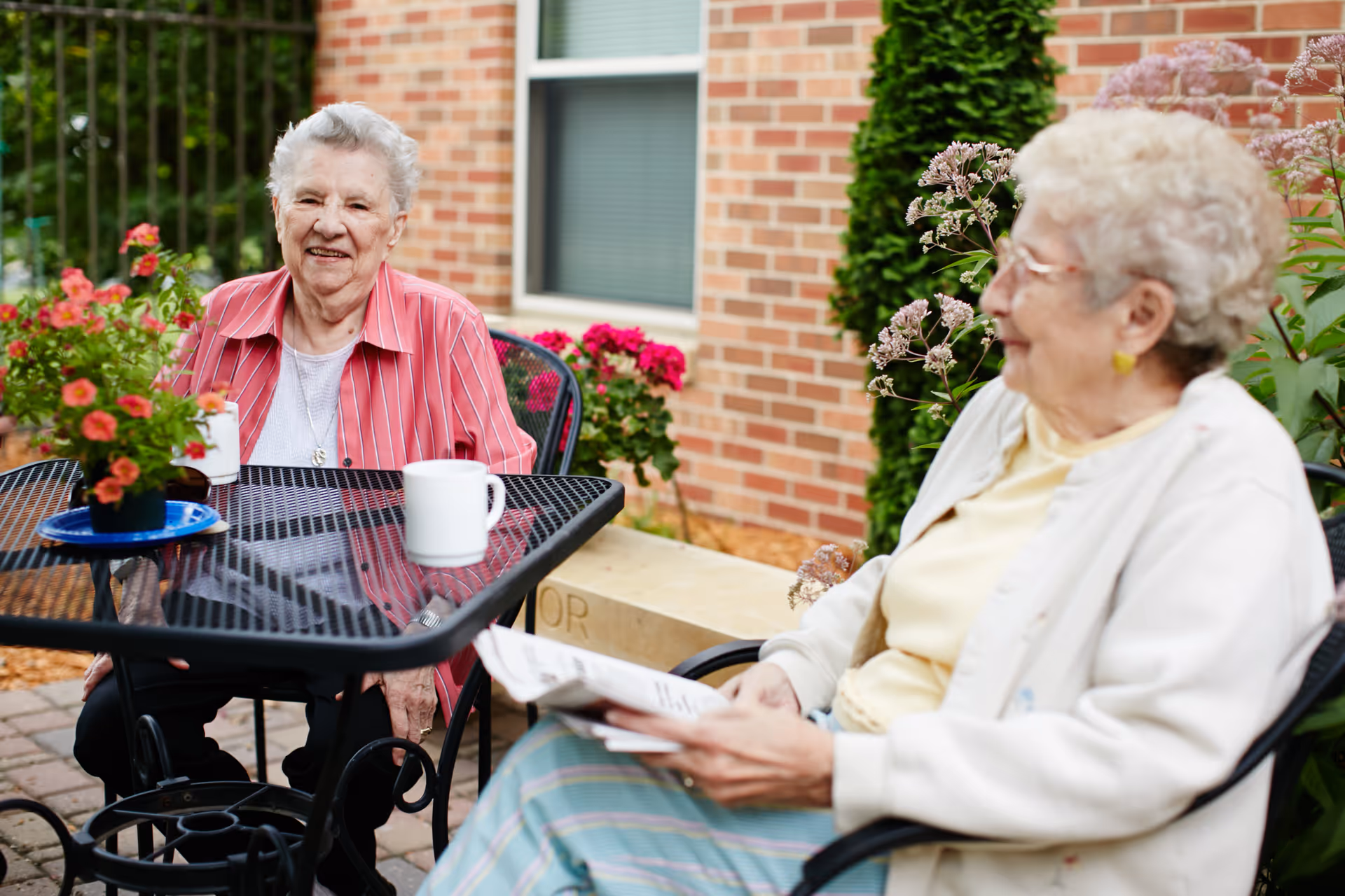 Two elderly women sitting outside at a black metal table with a potted plant and white mugs. One woman is wearing a red striped shirt and smiling at the camera, while the other woman is wearing a light-colored cardigan and holding a newspaper. They are seated in a garden area with flowers and a brick building in the background.