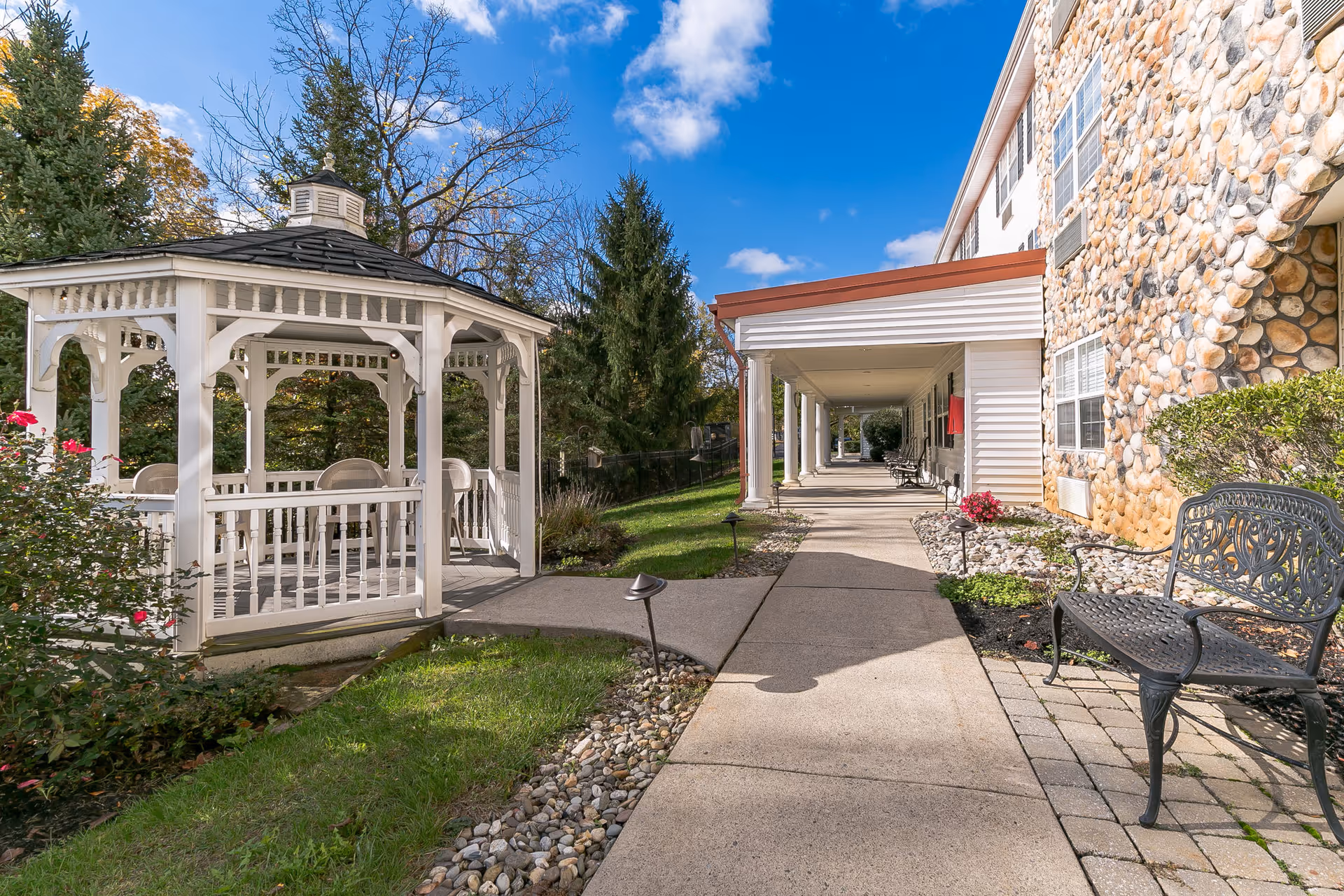 A paved walkway beside a senior living building with a white gazebo, benches, and landscaping under a blue sky.