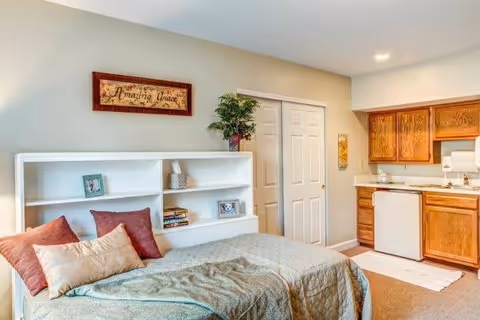 A cozy room featuring a bed with pillows and a quilted blanket, a white bookshelf with framed photos and books, a wall sign that reads 'Amazing Grace', and a kitchenette area with wooden cabinets, a sink, and a small refrigerator.