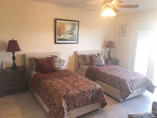 A bedroom with two twin beds side by side, each with patterned maroon and beige bedding and multiple pillows. There are two dark wooden nightstands with matching red lamps on either side of the beds. A ceiling fan with lights is on the ceiling, and a framed picture hangs on the wall above the beds. A door with glass panels is visible on the right side of the room.
